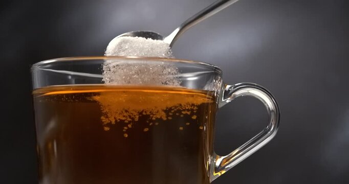 Sugar falling from spoon into hot black tea in glass cup in slow motion