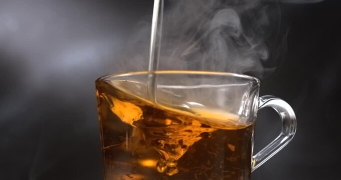 Hot black tea stirring with spoon in glass cup with steam on black background in slow motion