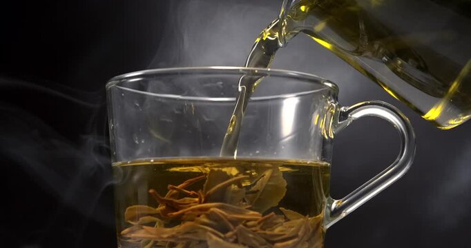 Hot green tea pouring into glass cup with tea leaves and steam in slow motion