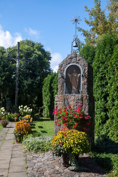 Vilnuis, Lithuania, 3 September 2026,  Stone wayside shrine with statue of Jesus, cross, bell, and potted flowers in churchyard. The Roman Catholic Church of St. Bartholomew in Uzupis district,