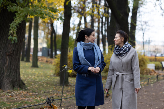 Mobility. Female friends strolling outdoors in cool season urban environment wearing coats and scarves communicating while moving through park area.