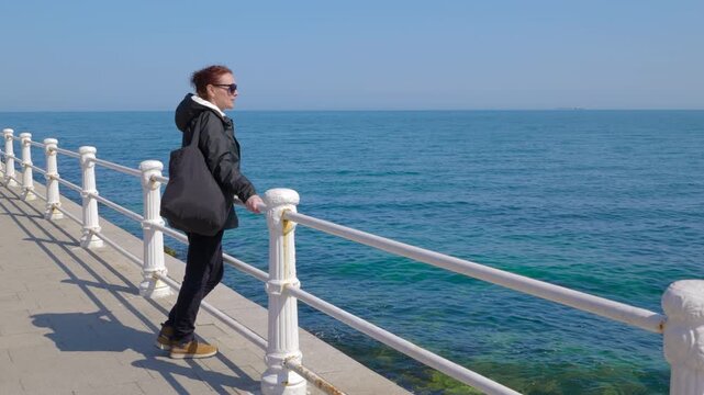 Woman admiring the ocean view from a seaside promenade. Young woman with red hair wearing sunglasses standing on a seaside promenade on a sunny day, looking at the calm blue sea