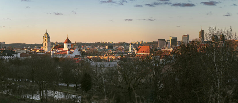 Panoramic view of Vilnius Old Town and city skyline in warm winter evening light. Historic church towers and red rooftops glow at sunset above a quiet urban landscape.