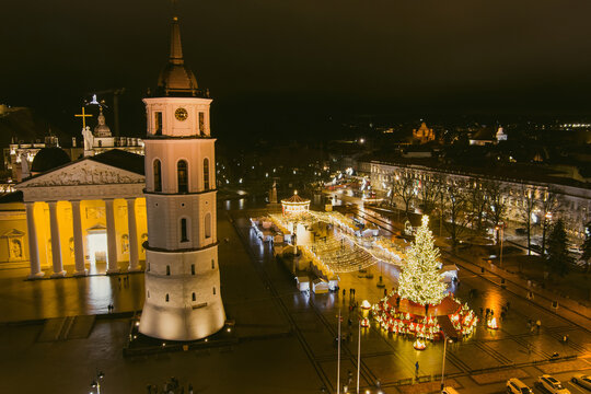 Aerial night view of the 2026 Christmas tree and festive market in Cathedral Square, Vilnius, Lithuania. Illuminated Vilnius Cathedral and Bell Tower glow beside holiday decorations.