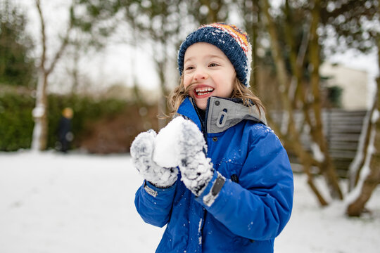Happy young child playing with fresh snow in the backyard on a winter day. Smiling kid holding snow in gloved hands and enjoying outdoor seasonal activity.