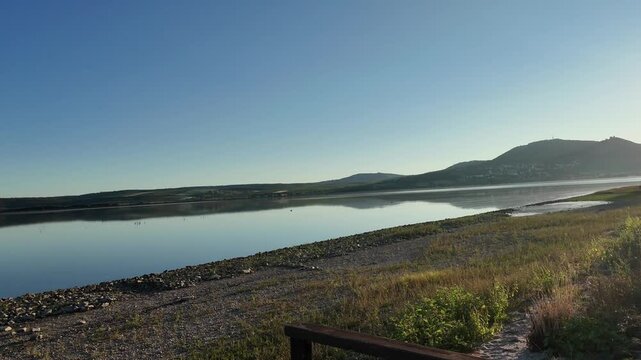 A serene view of a calm lake with a small fishing boat floating on the water at golden hour. The tranquil landscape features a rocky shoreline and distant hills under a clear blue sky while a lone fis
