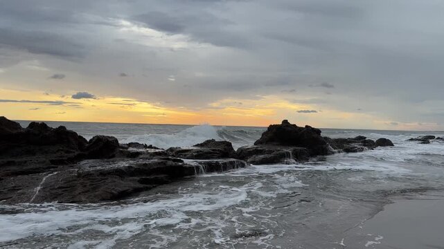The sun sets over the ocean as waves hit the rocky shore at the beach. The sky is filled with clouds and colors.