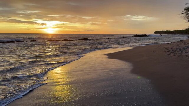 The sun sets over the beach while waves softly crash on the shore. The sky is filled with clouds and warm colors.