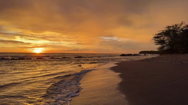 The sun sets on the horizon at a beach with calm waves moving ashore and a few small rocks in the water.