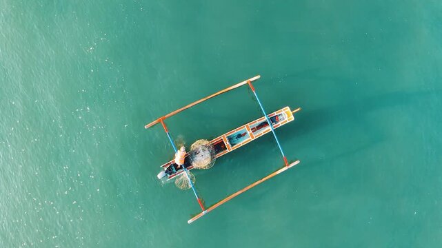 Top down aerial drone of Balinese fisherman casting fishing net from traditional wooden jukung outrigger boat on clear turquoise sea. Splash water action Yeh Gangga Bali Indonesia