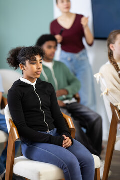 Thoughtful teenage girl listening to teacher while sitting with classmates in modern school classroom