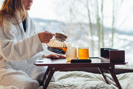 Drinking hot tea enjoying winter landscape in mountains. Woman wears bathrobe relaxing by big window of cabin house on holiday