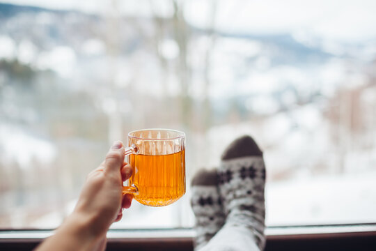 Drinking hot tea enjoying winter landscape in mountains. Woman wears warm socks relaxing by big window of cabin house on holiday