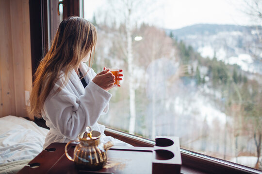 Drinking hot tea enjoying winter landscape in mountains. Woman wears bathrobe relaxing by big window of cabin house on holiday