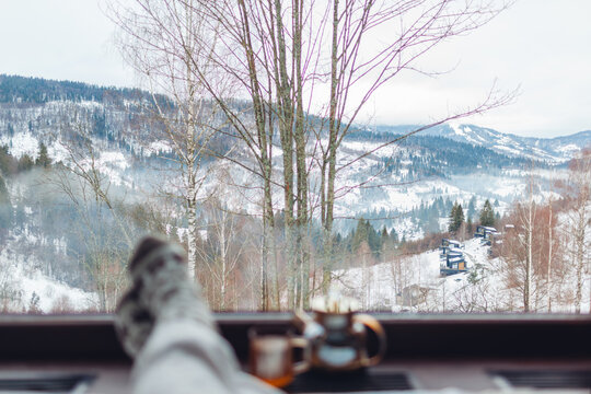 Drinking hot tea enjoying winter landscape in mountains. Woman wears warm socks relaxing by big window of cabin house on holiday