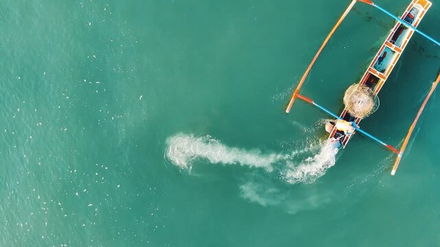 Top down aerial drone view of fishing net sinking with white wake trail on turquoise sea water from traditional jukung outrigger boat. Balinese fisherman fishing action Yeh Gangga Bali