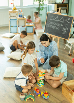 Toys are scattered on floor, children playing with cubes and cars, babysitter support and participate in game. Kindergarten or childcare center or daycare, classes for kids.