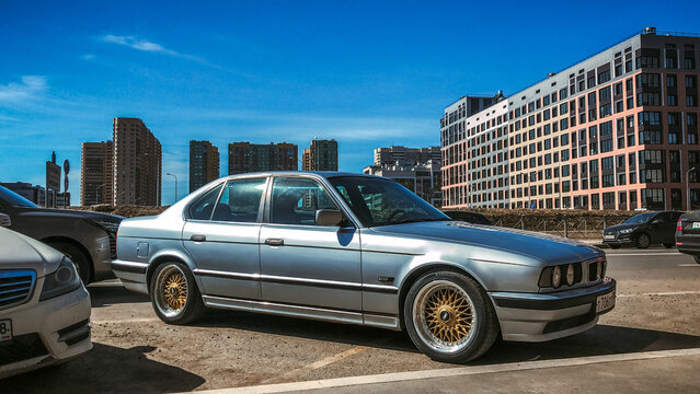 Classic silver BMW 5 Series E34 German retro car parked on a city street close up. Famous sedan passenger automobile with custom golden alloy wheels near modern multi story apartment buildings. 