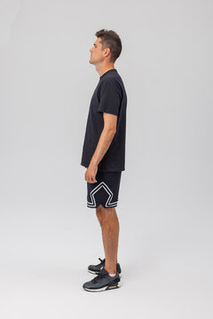 Full profile view of an athletic young man standing against a gray studio background, looking forward. He is wearing a blank black t-shirt, geometric patterned sports shorts, and modern sneakers.