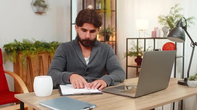 Indian man at home reviews paper reports compares numbers with laptop writes notes focused. Arabian guy at desk checks rows and figures on sheet reads data finds match does careful analysis calm mood