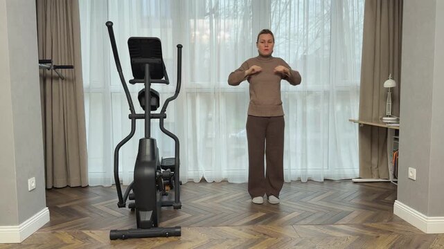  Woman wearing warm knitted suit and sneakers performs shoulder activation, bringing arms forward and squeezing shoulder blades in cozy living room.