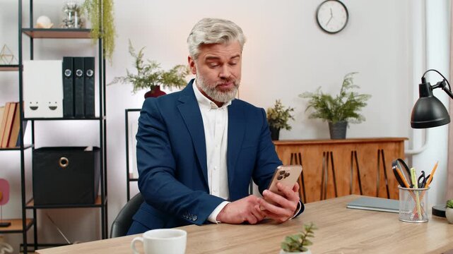 Middle-aged businessman at home office stares at smartphone reading project failure notice breathes deep feeling despair. Freelancer guy at table sighs sadly shoulders slumped planning next work step.