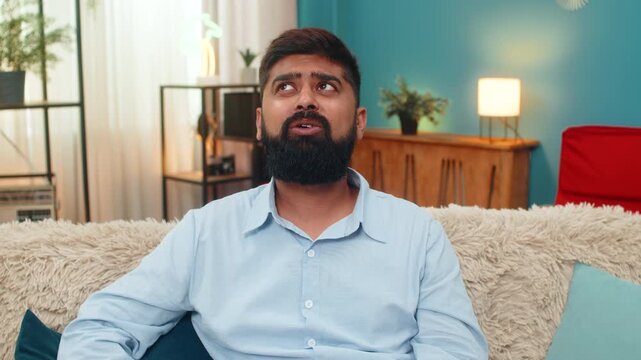 Indian man at home rolls eyes showing ironic reaction during smartphone chat dismissing odd comment. Arabian guy on sofa shows sarcasm disbelief slight head tilt expresses annoyance humorous attitude.