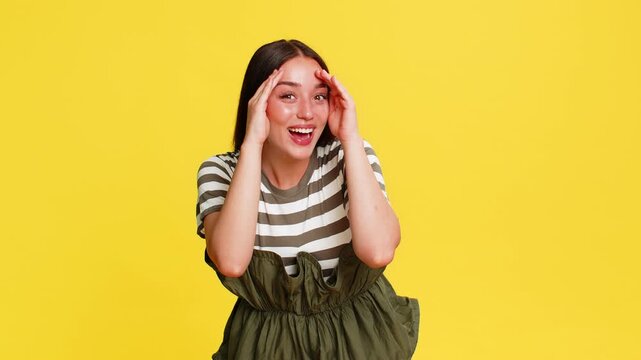 Young woman hiding face behind hands and then peeking out with gentle smile, expressing bashful shyness. Girl on yellow background shows playful emotional reaction of modesty and light embarrassment.