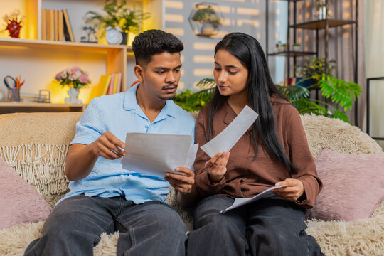 Indian couple man and woman sitting on home sofa check bills and receipts for budget planning. Young family man and woman sitting on home sofa count expenses and discuss utility payments each month.