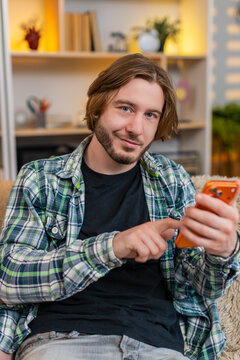 Young man on home sofa types quick smartphone message then stops to reread words with focused smile. Adult guy on home sofa presses send with calm satisfaction after checking text and finishing chat.