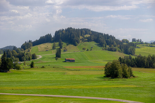 Rural Bavarian countryside landscape, Traditional wooden houses and barn on rolling hills, Lush green grass meadow against majestic Alpine mountains under blue sky and white clouds in Bavaria, Germany