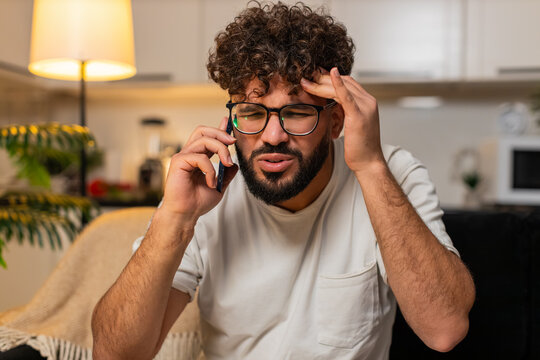 Arabian man at home answers smartphone gets spam call from pushy operator says no feels angry annoyed. Egyptian guy on sofa rejects fake offer blocks number sighs frowns frustrated after fraud call.