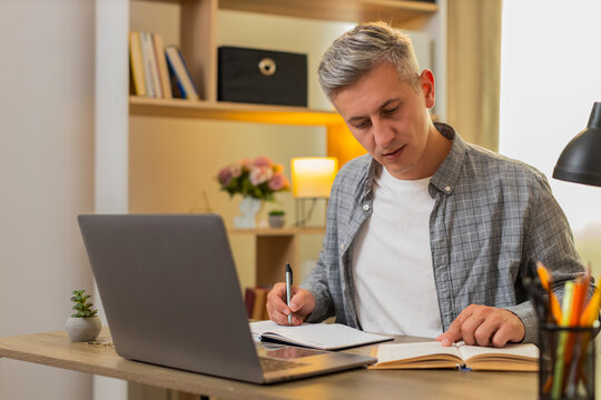 Middle-aged man on home table compares book text with laptop research and searches useful information. Young adult guy on home table checks facts between sources and writes notes for focused studies.