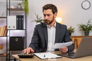 Middle-aged businessman at home office enters numbers on laptop checks amounts with calculator and...