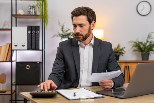 Middle-aged businessman at home office enters numbers on laptop checks amounts with calculator and receipts. Freelancer guy at table reviews bills and signs documents with calm focus and steady hand.