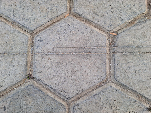 Gray hexagonal concrete paving blocks arranged in interlocking pattern, showing rough texture, natural wear, cracks, and small weeds growing in gaps