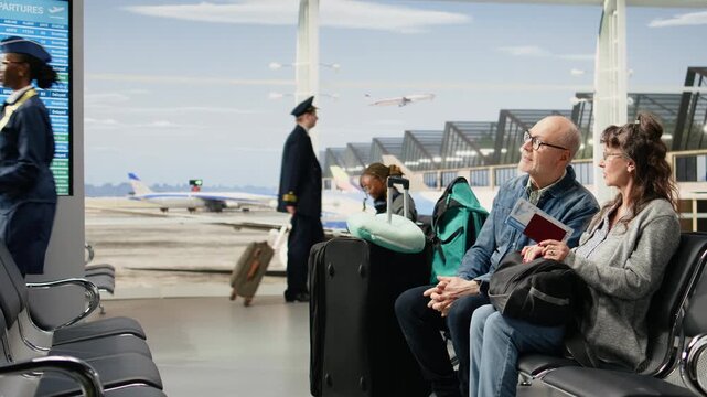 Smiling elderly couple sits in an airport lounge with luggage, chatting and planning their trip while waiting for the plane, embracing retirement freedom, bucket list travel goals, and later life.