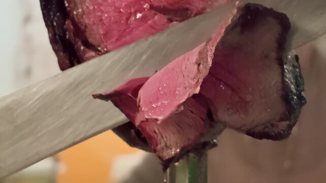 Closeup of large sharp knife slicing juicy picanha roast beef from a metal skewer, revealing perfectly cooked pink interior at a traditional Brazilian churrascaria rodizio service. Elegant lifestyle.