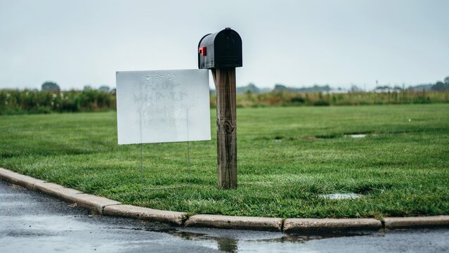 Blank white yard sign standing on a post beside a black mailbox in a green roadside lawn