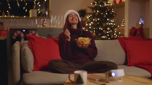 Woman in Santa hat eating chips while watching comedy movie at home