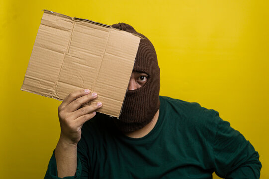 Young Asian male wearing brown balaclava holds blank corrugated board over one eye against bright yellow backdrop. The masked individual wears dark green apparel, creating striking