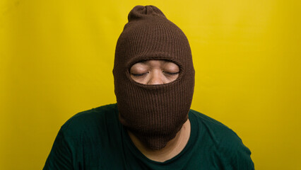 Young Asian male wearing brown balaclava stands with closed eyes against vivid yellow backdrop. The masked individual projects an aura of peacefulness, resignation, or deep concentration, © M. Faisal Riza