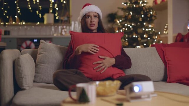 Scared woman in Santa hat hugging pillow while watching movie at home