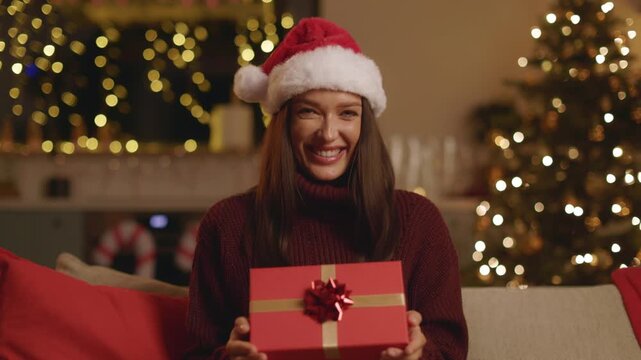 Smiling woman in Santa hat holding wrapped Christmas gift at home, 4K