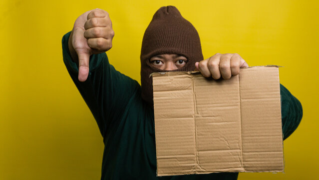Young Asian male wearing brown balaclava holds blank cardboard sign while displaying negative thumbs down gesture against bright yellow background. The masked figure expresses disapproval