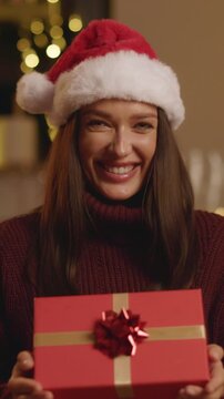 Smiling young woman in Santa hat holding wrapped Christmas gift, vertical 4K