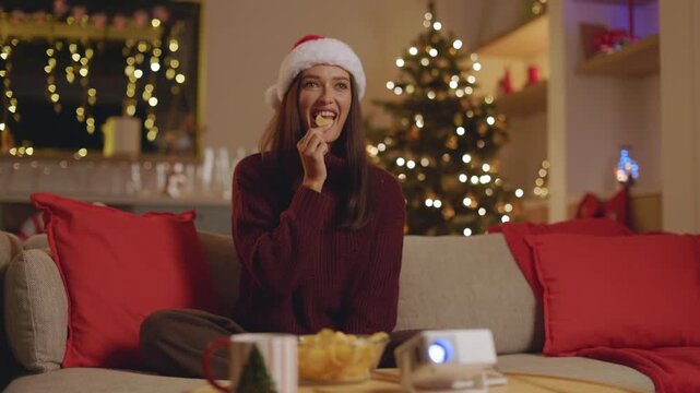 Woman in Santa hat reaching for chips while enjoying projector movie, 4K