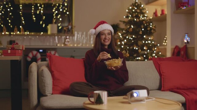 Smiling young woman in Santa hat enjoying chips and watching movie, 4K