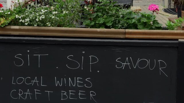Chalkboard sign beside lush flower planter with handwritten sit sip savour local wines craft beer message