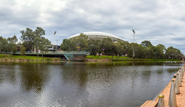 The River Torrens flows through Adelaide, South Australia, with the modern Riverbank Precinct Pedestrian Bridge leading toward the iconic Adelaide Oval stadium